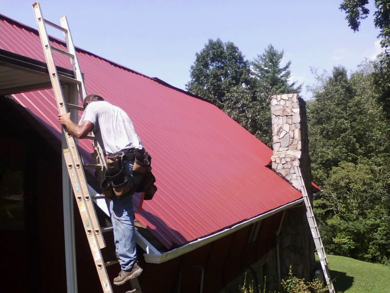 Roofer on a ladder beside a red metal roof and stone chimney — Soffit & Fascia Repair by Roofing Brothers in Coeymans